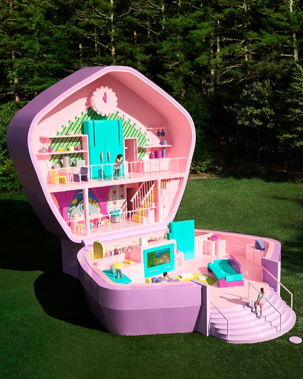 Uma casa de bonecas gigante com um design moderno e colorido. A decoração interna inclui móveis vibrantes em tons de rosa, lavanda e turquesa. As duas bonecas vestem roupas casuais e coloridas, seguindo tendências infantis de moda.