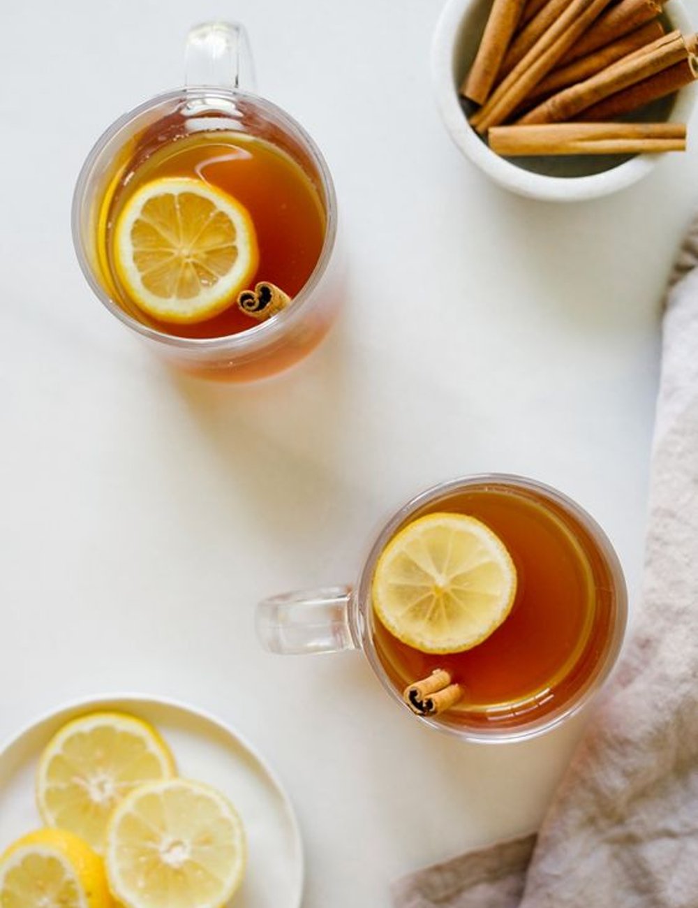 Imagem de duas canecas de vidro com chá de canela e limão, acompanhadas de um prato com fatias de limão e um pote com paus de canela. Estilo minimalista e tendência de bebidas saudáveis e naturais. Soprar canela.