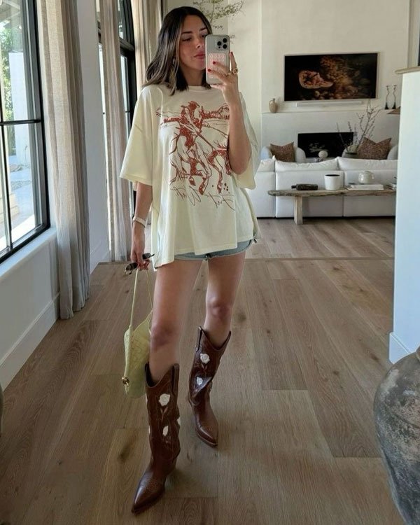 Mulher vestindo camiseta oversized com estampa artística, shorts jeans curtos e botas cowboy marrons com detalhes. Look moderno e casual, combinando conforto com elementos do estilo country urbano.