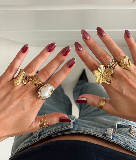 Unhas vermelhas decoradas com acabamento ovalado, várias anéis grandes e dourados com pedras e desenhos florais, vestindo jeans de cintura baixa e top preto. tendência boho e acessórios chamativos.