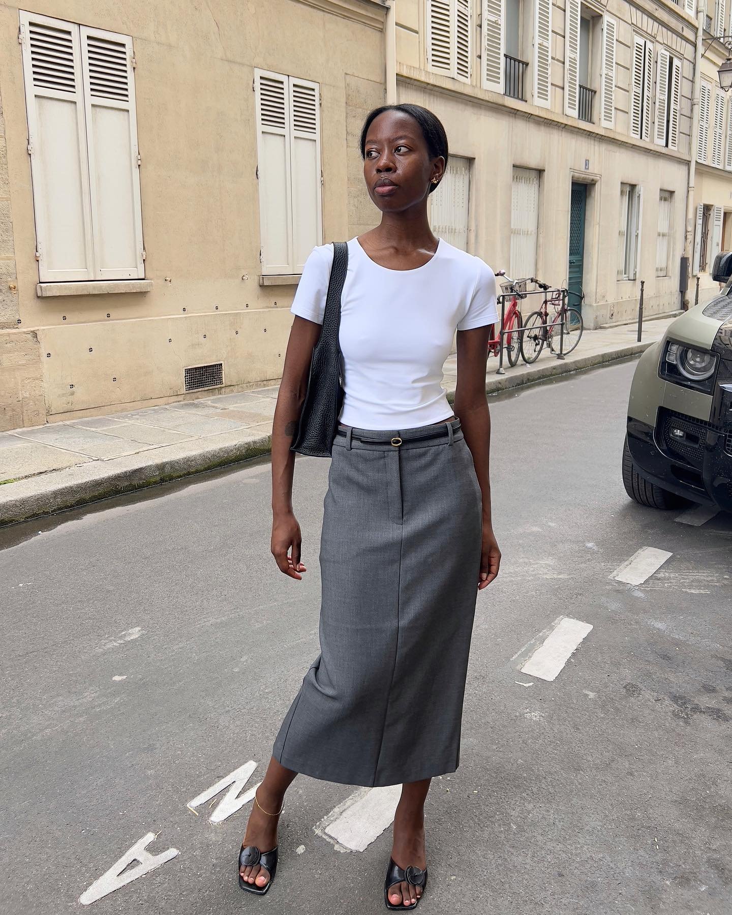 Mulher veste camiseta branca básica, saia midi cinza de cintura alta e sapatos abertos pretos com salto. O look é complementado por uma bolsa preta e uma tornozeleira dourada.