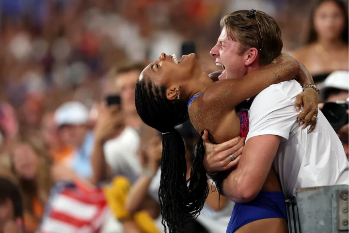 Mulher em roupa esportiva azul e vermelha abraça homem de camiseta branca em meio a uma multidão. Ela usa tranças longas e molhadas, destacando estilo vibrante e casual, típicos de eventos esportivos. Cena de celebração e emoção.