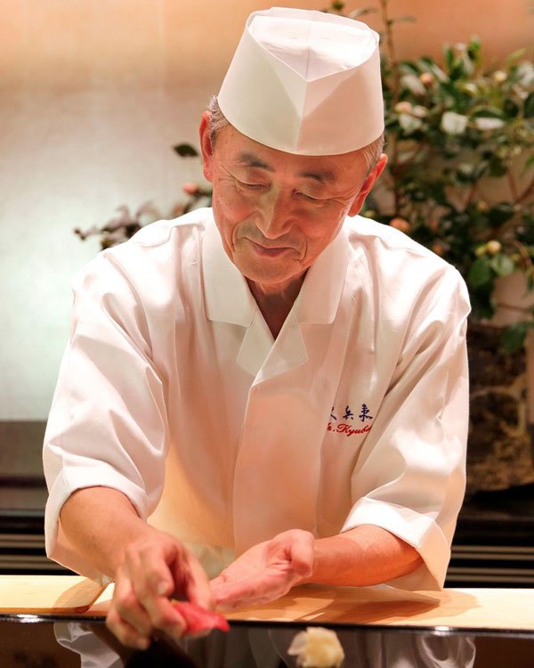 Chef de sushi vestido com uniforme tradicional japonês, incluindo um casaco branco de chef com botões e um chapéu toque blanche. A vestimenta reflete a culinária de alta classe e segue tendências de elegância e simplicidade na apresentação pessoal.