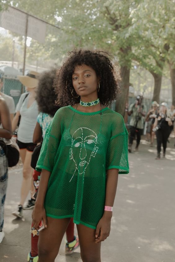 Pessoa vestindo camiseta verde de malha com estampa de rosto estilizado, combinada com shorts curtos. Acessórios incluem choker verde, brincos grandes e anéis. Estilo casual e urbano, perfeito para festivais ao ar livre e eventos de verão.