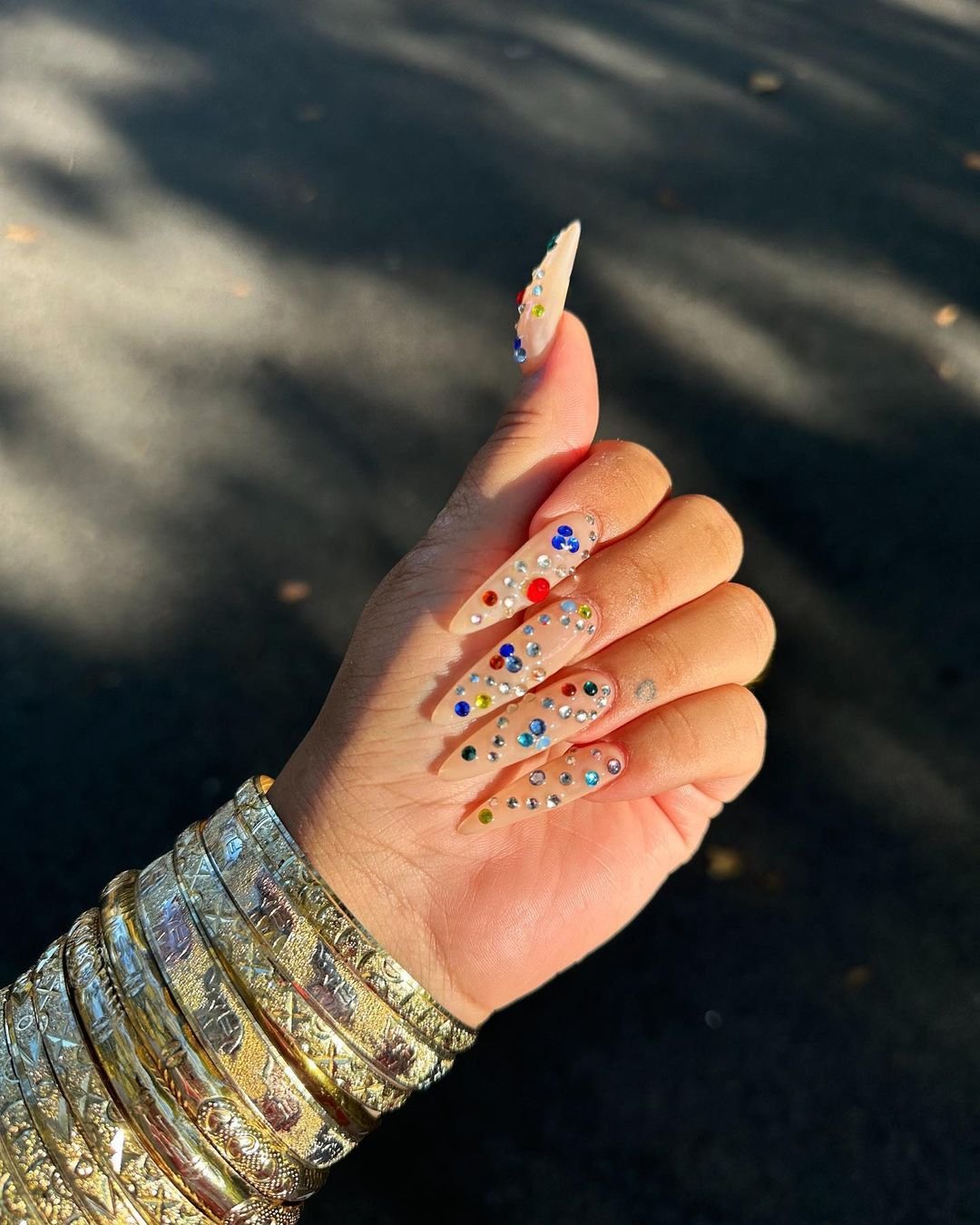  tendências de nail art 
 Unhas postiças longas com decoração colorida de pedras brilhantes e pulseiras douradas detalhadas em camadas. Look moderno que combina criatividade nas unhas com acessórios tradicionais. Ideal para quem busca estilo sofisticado e tendência.