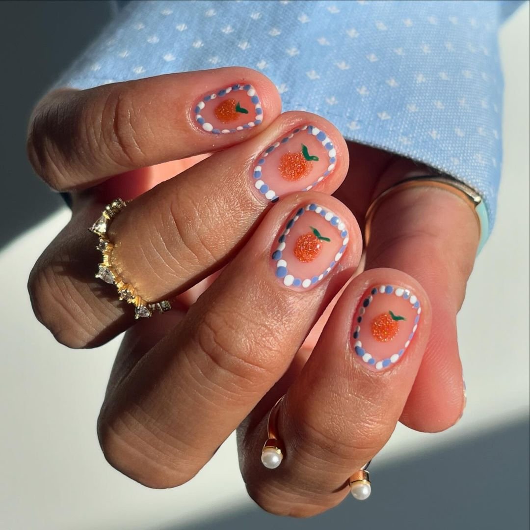 Unhas decoradas com detalhes de frutas e borda de bolinhas, seguindo a tendência de nail art minimalista. As mãos ainda exibem anéis delicados e elegantes, destacando um apelo fashion moderno e sutil. tendências de nail art