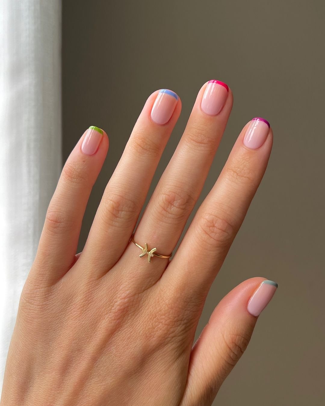 tendências de nail art curtas com esmaltação francesa colorida, cada ponta em uma cor diferente, tendência moderna e minimalista. Anel delicado de ouro no dedo anelar adiciona um toque elegante ao visual das mãos.