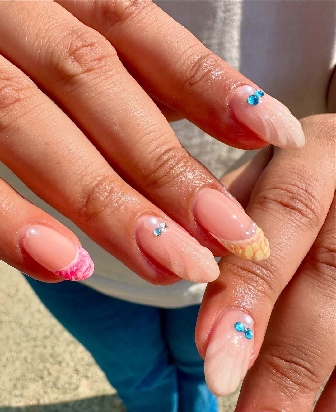 Unhas decoradas com tendências de nail art elegante, combinando tons de nude e detalhes em rosa com pedrarias azuis. As unhas apresentam diferentes padrões e texturas, ressaltando uma tendência moderna e artística para manicure.