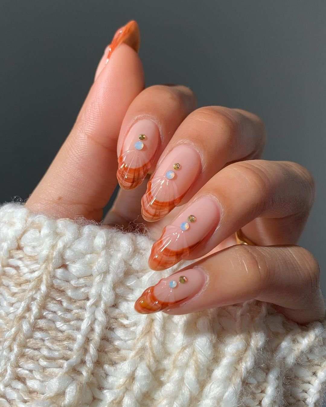 tendências de nail art com esmalte em tons de laranja e detalhes marmorizados, acompanhadas por pequenas joias. A mão está vestindo um suéter de lã branco, criando um visual elegante e moderno, ideal para o outono e inverno. Tendência atual em nail art.