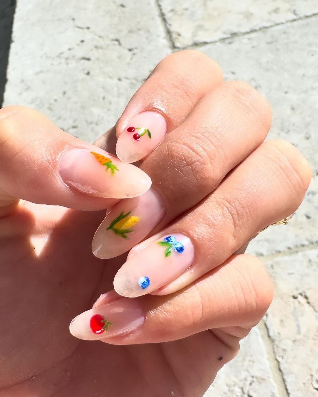 Unhas decoradas com tendências de nail art delicada e minimalista, apresentando desenhos de flores e frutas. A tendência destaca o estilo natural e colorido, perfeito para um visual moderno e descontraído. Ideal para quem busca inovação e criatividade nas unhas.