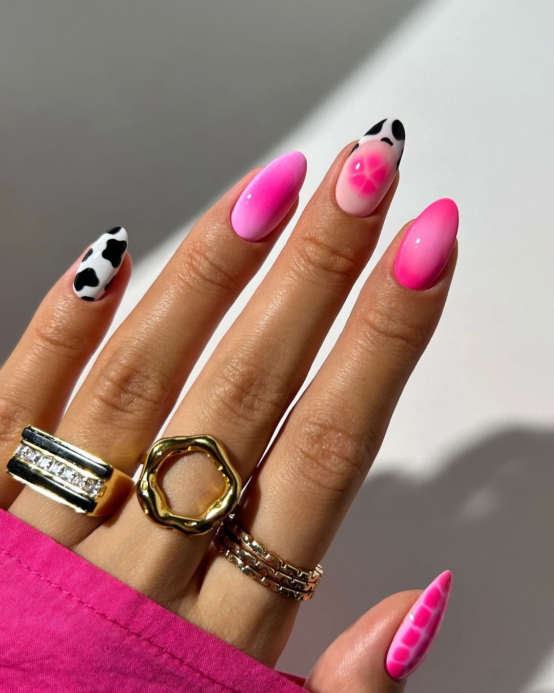 tendências de nail art em estilo moderno com mix de estampas de vaquinha e degradê rosa. Anéis dourados de diferentes formatos complementam o look. Tendência de moda em nail art e acessórios elegantes. Blusa rosa dá destaque ao visual.