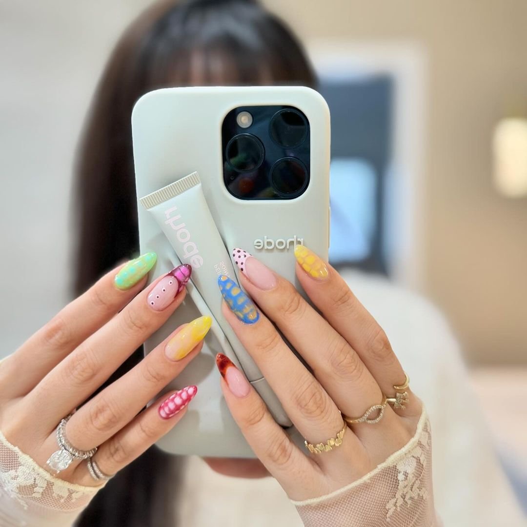 tendências de nail art em tons vibrantes apresentam desenhos variados, enquanto a pessoa segura um smartphone. Anéis dourados complementam o visual, que inclui uma manga com renda delicada, destacando um estilo moderno e sofisticado.