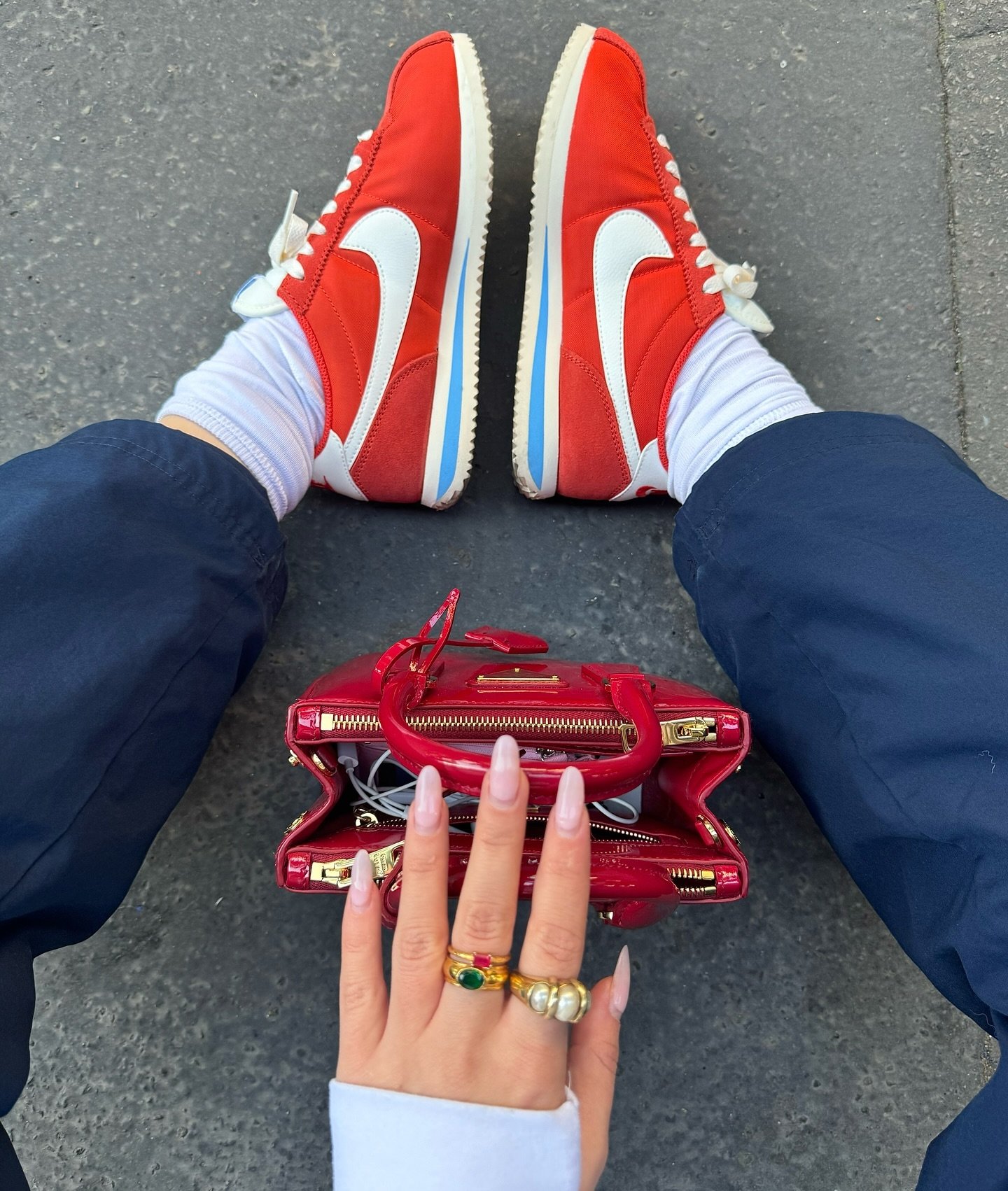 Tênis Nike vermelhos combinados com calça esportiva azul e meias brancas. Bolsa vermelha e vários anéis dourados chamativos. Unhas longas e bem cuidadas. Estilo casual e urbano, com forte influência streetwear. Acessórios elegantes destacam o visual.