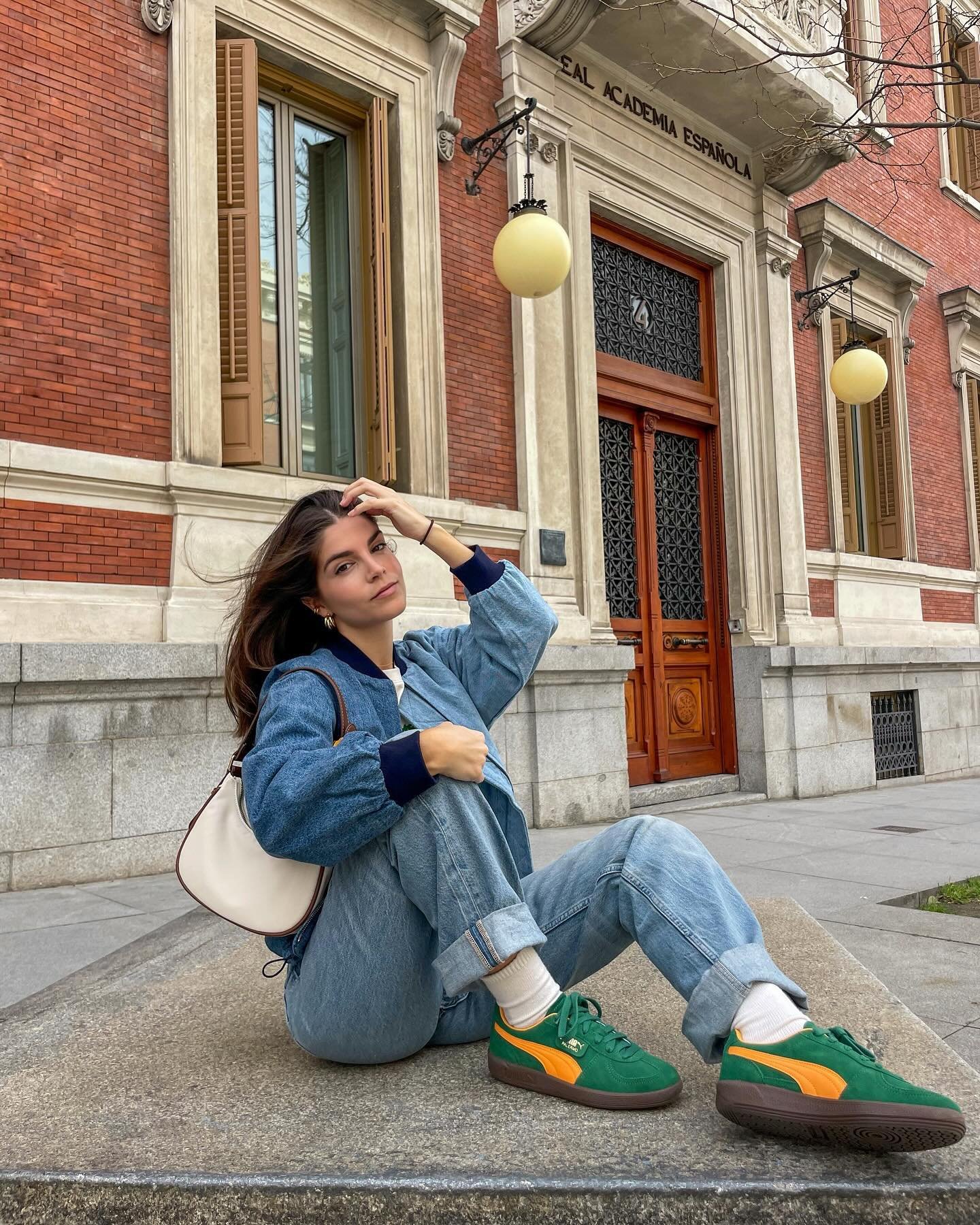 Jovem usando conjunto jeans oversized (calça e jaqueta), camiseta branca e tênis verde com detalhes amarelos. Complementa o look com bolsa pequena. Estilo casual e despojado, perfeito para passeios urbanos. Fundo com edificação clássica em tijolos aparentes.