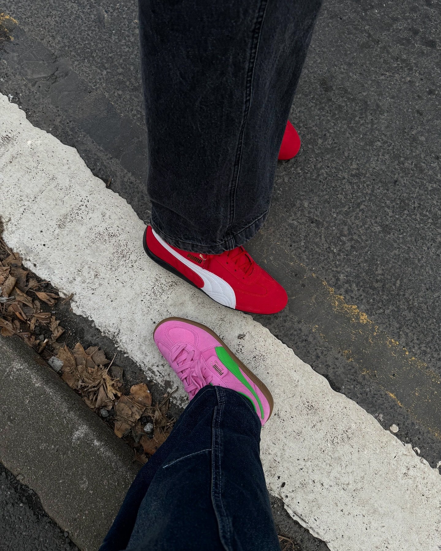 Dois pares de tênis Puma em destaque na imagem: um par rosa com detalhes verdes e um par vermelho com detalhes brancos. A moda urbana e casual se destaca, com ambos vestindo jeans escuros. Tendências de tênis coloridos e estilos streetwear.