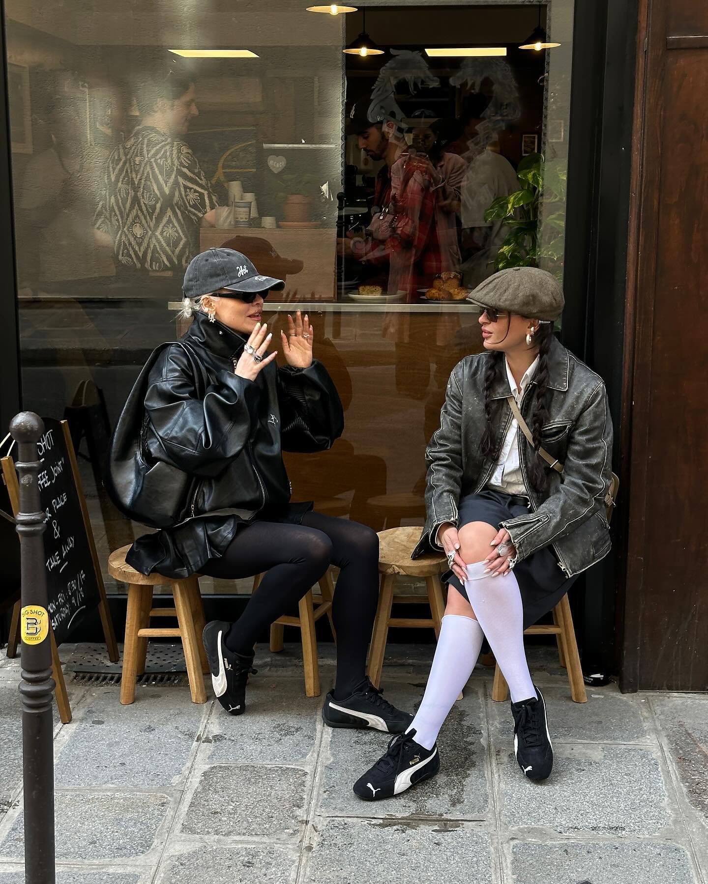 Duas mulheres sentadas em frente a um café, ambas usando bonés e roupas esportivas. Uma veste jaqueta e calça legging preta, a outra jaqueta cinza, bermuda preta e meias compridas brancas. Ambas calçando tênis pretos. Estilo urbano e casual.