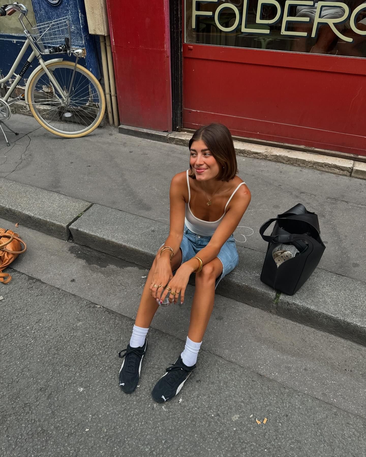 Pessoa sentada na calçada usando regata branca, saia jeans e tênis preto com meias brancas. Acessórios incluem diversas pulseiras e anéis dourados. O estilo é casual chic, ideal para um dia descontraído na cidade.