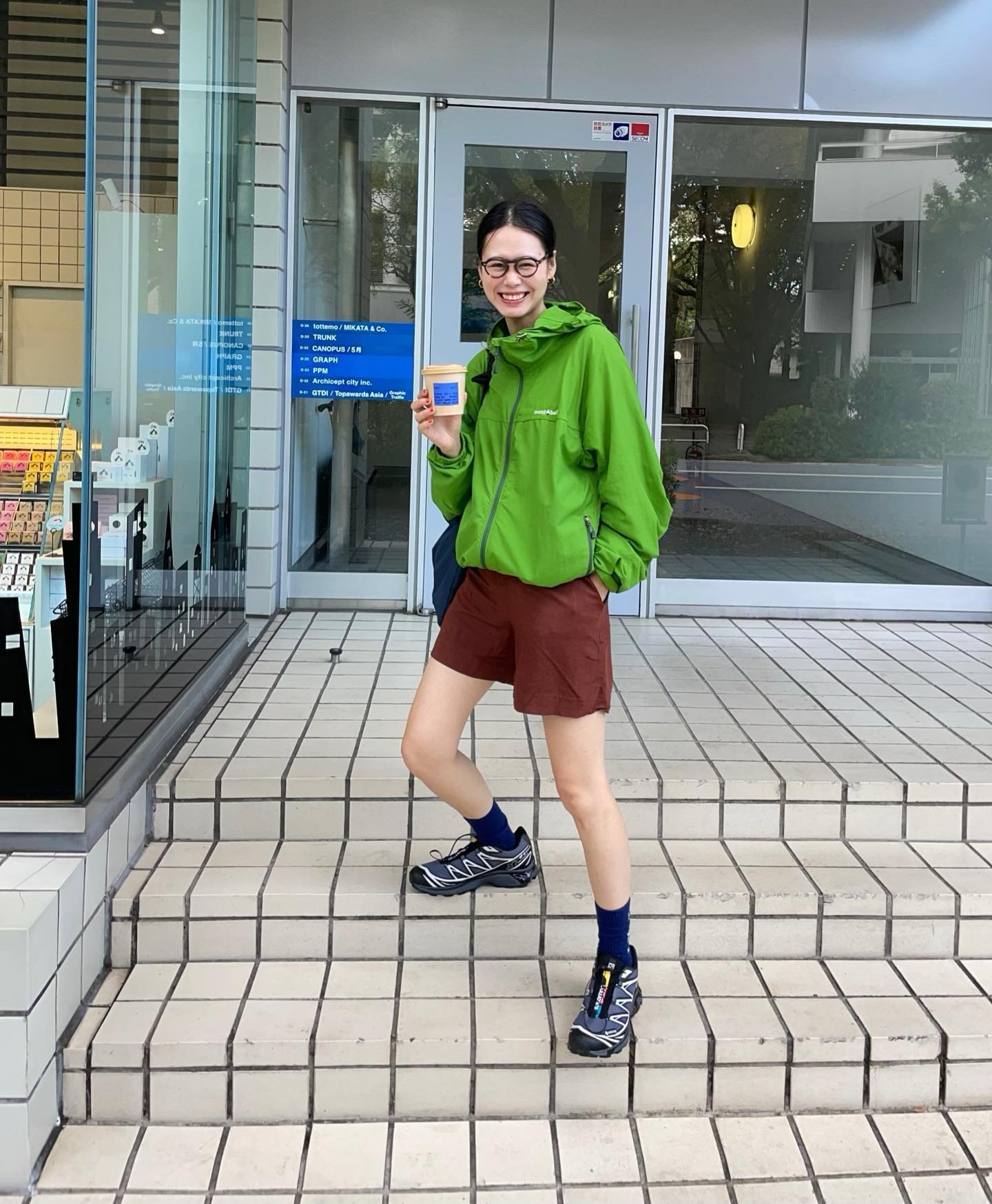 Pessoa vestindo jaqueta verde, shorts marrom, meias azuis e tênis esportivos, posando em frente a uma loja moderna. Estilo casual e esportivo com tendência urbana.