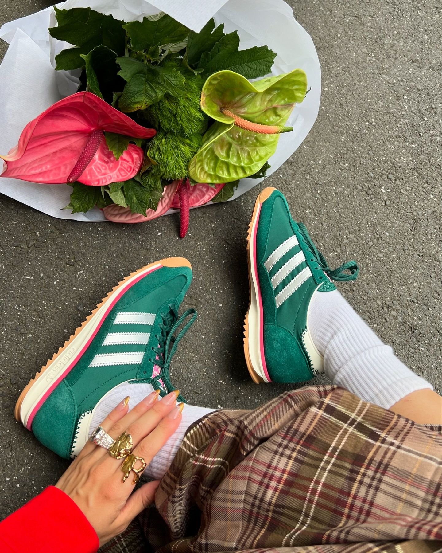 Tênis verde com listras brancas da Adidas combinados com meias brancas e saia xadrez em tons terrosos. Look complementado com anéis dourados e unhas longas estilo nail art, destacando o visual moderno e descolado. Flores exóticas também estão presentes.