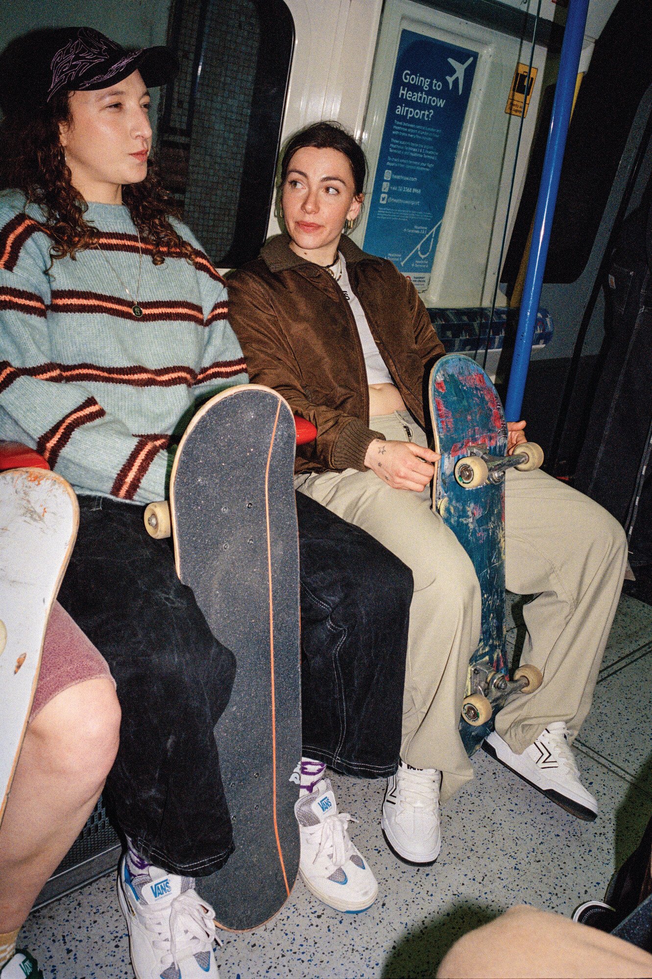 Duas jovens em um metrô segurando skates, vestem-se no estilo urbano e skatista: uma usa suéter listrado, calça preta larga e boné; a outra, jaqueta marrom, calça bege e cropped. Ambas calçam tênis casuais, ambas usando os tênis vans.