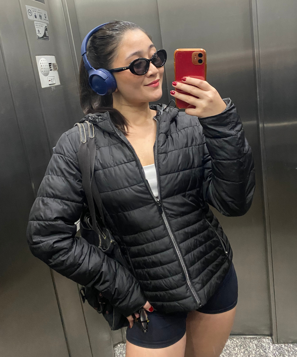 Modo esportivo casual: Jaqueta puffer preta, top branco, shorts biker preto, óculos de sol, fones de ouvido azuis e moletom. Look urbano confortável e prático, combinando peças básicas e tendências em um estilo casual e esportivo.