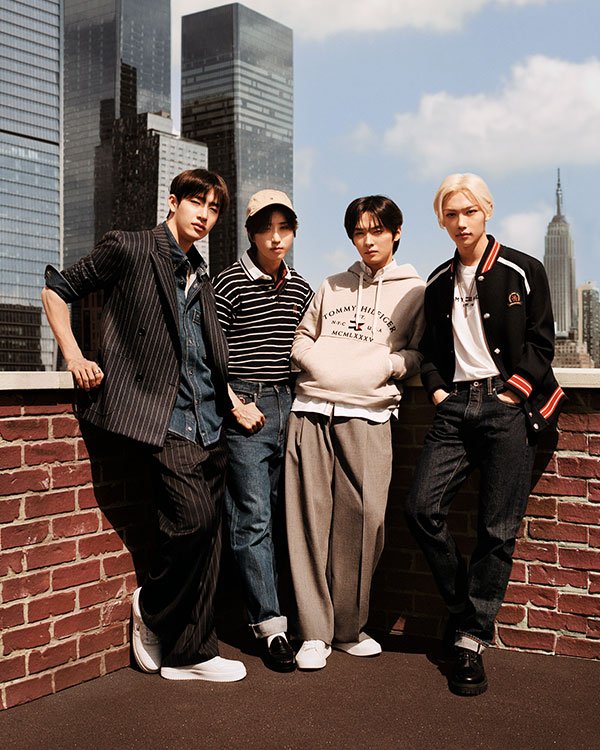 Quatro homens jovens posam em um rooftop vestindo tendências urbanas e modernas. Looks incluem ternos listrados com camiseta, suéter listrado com boné, moletom casual com calça social larga, e jaqueta desportiva combinada com jeans.