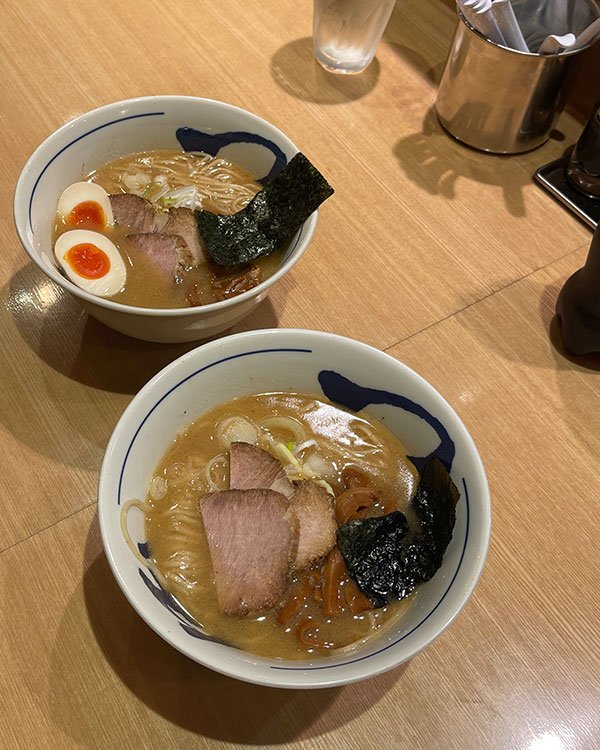 Duas tigelas de ramen em um balcão de madeira. O prato contém ovo cozido, nori, fatias de carne de porco, macarrão e caldo rico. A moda japonesa tradicional está presente na apresentação cuidadosa e minimalista do prato.