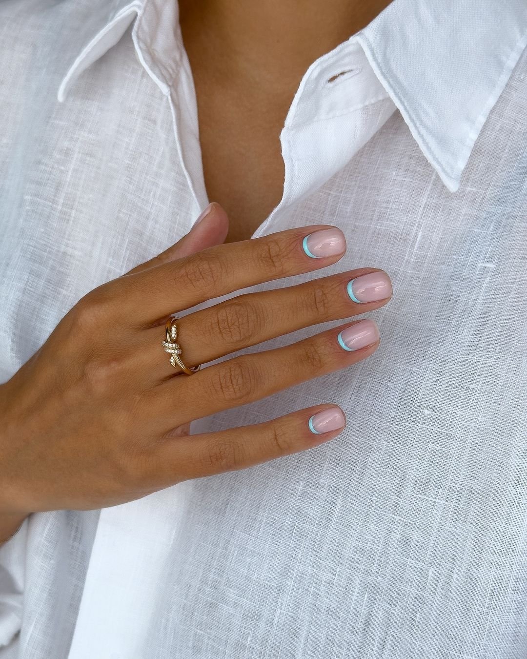 Camisa social branca de linho, unhas decoradas com esmalte nude e linhas finas em azul nas pontas, e anel dourado delicado. Visual elegante e minimalista, ideal para o dia a dia ou ocasiões formais, unhas encravadas