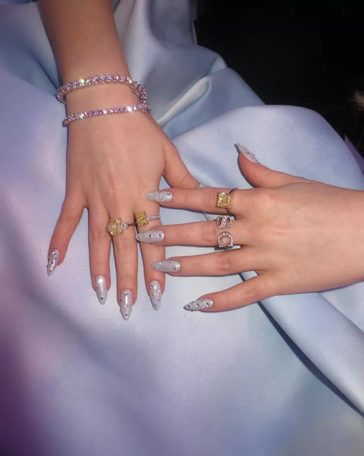 Unhas decoradas com esmalte prateado e pedrarias, acompanhadas por diversos anéis chamativos em tons de dourado e prateado. As pulseiras brilham com cristais, destacando um estilo luxuoso e moderno. Sabrina Carpenter