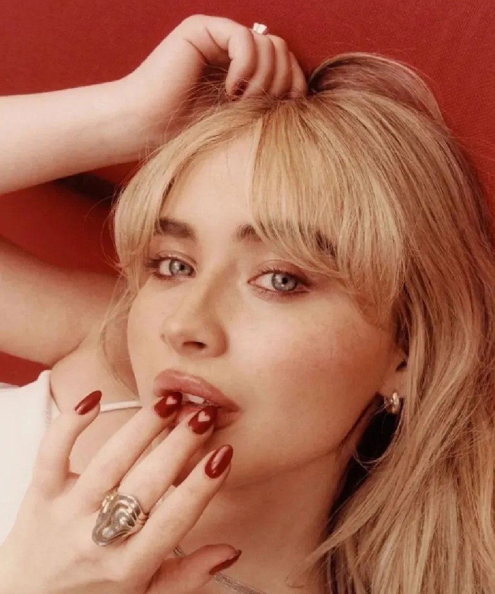 Imagem de uma mulher com cabelo loiro e franja, maquiagem suave e foco nas unhas esmaltadas de vermelho. Usa um anel grande e elegante, sugerindo tendências de moda moderna e minimalista. Sabrina Carpenter