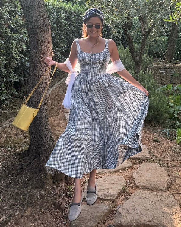 Mulher usando vestido midi xadrez, estilo clássico, com alças largas. Acessórios incluem bolsa amarela, óculos de sol redondos e faixa no cabelo. Sapatos fechados e uma echarpe branca completam o look.