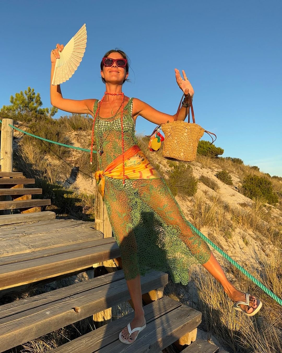 Mulher vestindo saída de praia verde rendada, por cima do biquíni, segura leque e bolsa de palha. Usa chinelos brancos e óculos de sol grandes. Tendências de moda incluem tecidos leves, acessórios coloridos e estilo boho praiano.