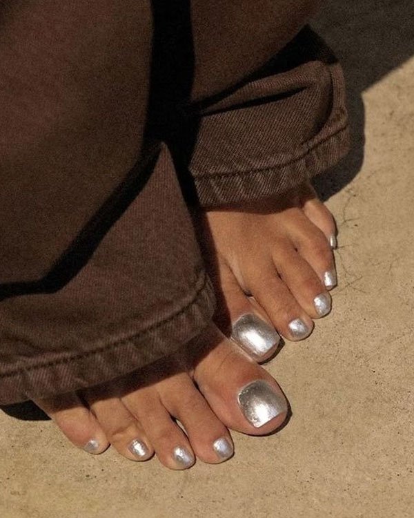 Unhas dos pés pintadas de prata metálico, combinadas com calças marrons de barra dobrada. Look casual e moderno, destacando a tendência de esmaltes metálicos e detalhes despojados em roupas.