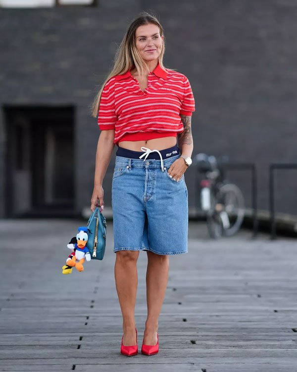 Mulher usando uma blusa polo cropped listrada vermelha, bermuda jeans folgada e tênis vermelhos. Segura uma bolsa azul com um chaveiro do Pato Donald.