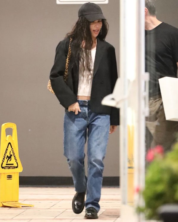 Pessoa vestindo tendências casuais e modernas: boné preto, blazer preto oversized, cropped branco, jeans azuis de corte reto e mocassim preto. Bolsa de ombro com estampa animal print. Estilo despojado e contemporâneo.