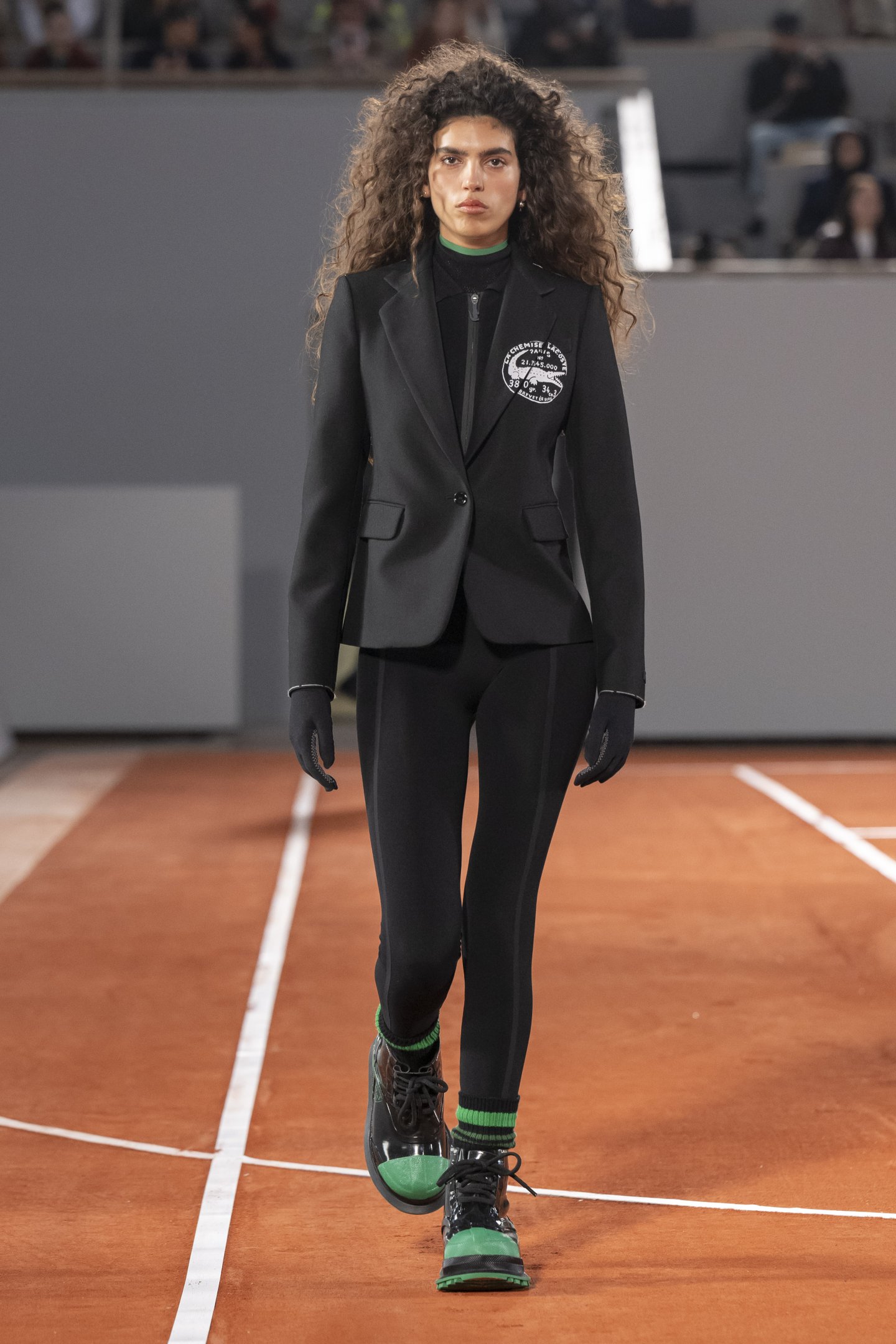 Modelo usa conjunto preto esportivo com toque elegante: blazer ajustado com emblema branco, blusa de gola alta, calça justa, luvas e botas verde e preta. Combinação de moda urbana e athleisure, destacando conforto e sofisticação.