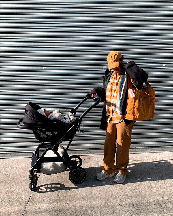 Pessoa usando looks casuais de inverno/entre-safra; camisa xadrez em tons terrosos, calça solta, boné e suéter preto sobre os ombros. Carrega bolsa grande e empurra carrinho de bebê. Moda prática e confortável para o dia a dia urbano.