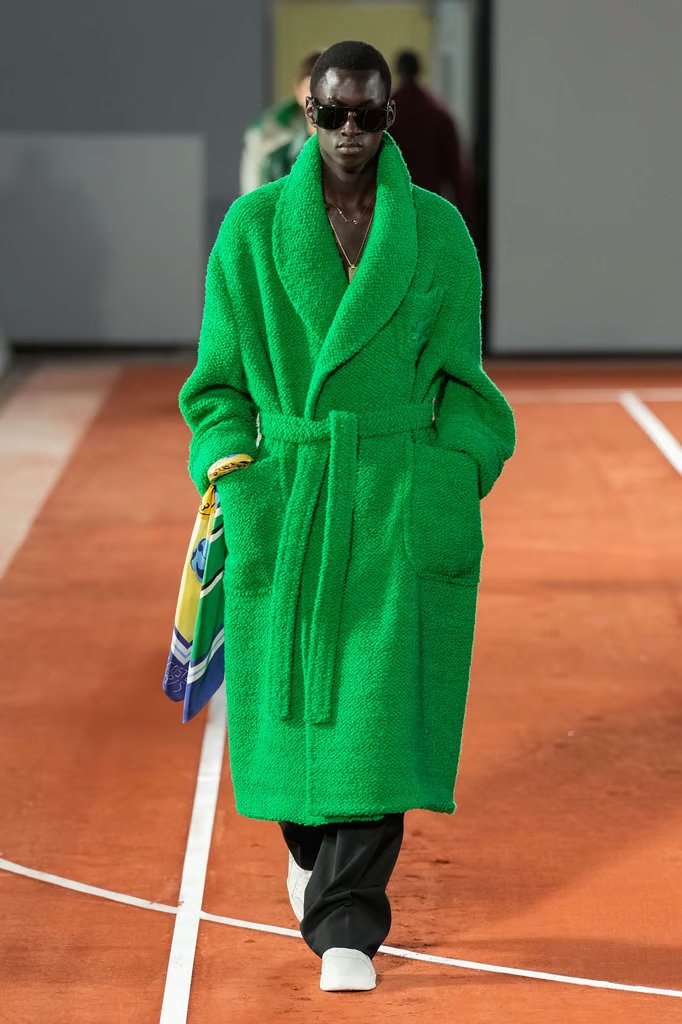 Desfile de moda com um casaco verde texturizado, estilo oversized, que remete a um roupão. A peça é combinada com calça preta, tênis brancos e óculos de sol grandes. Tendências: oversized, texturas marcantes, cores vibrantes e acessórios chamativos.