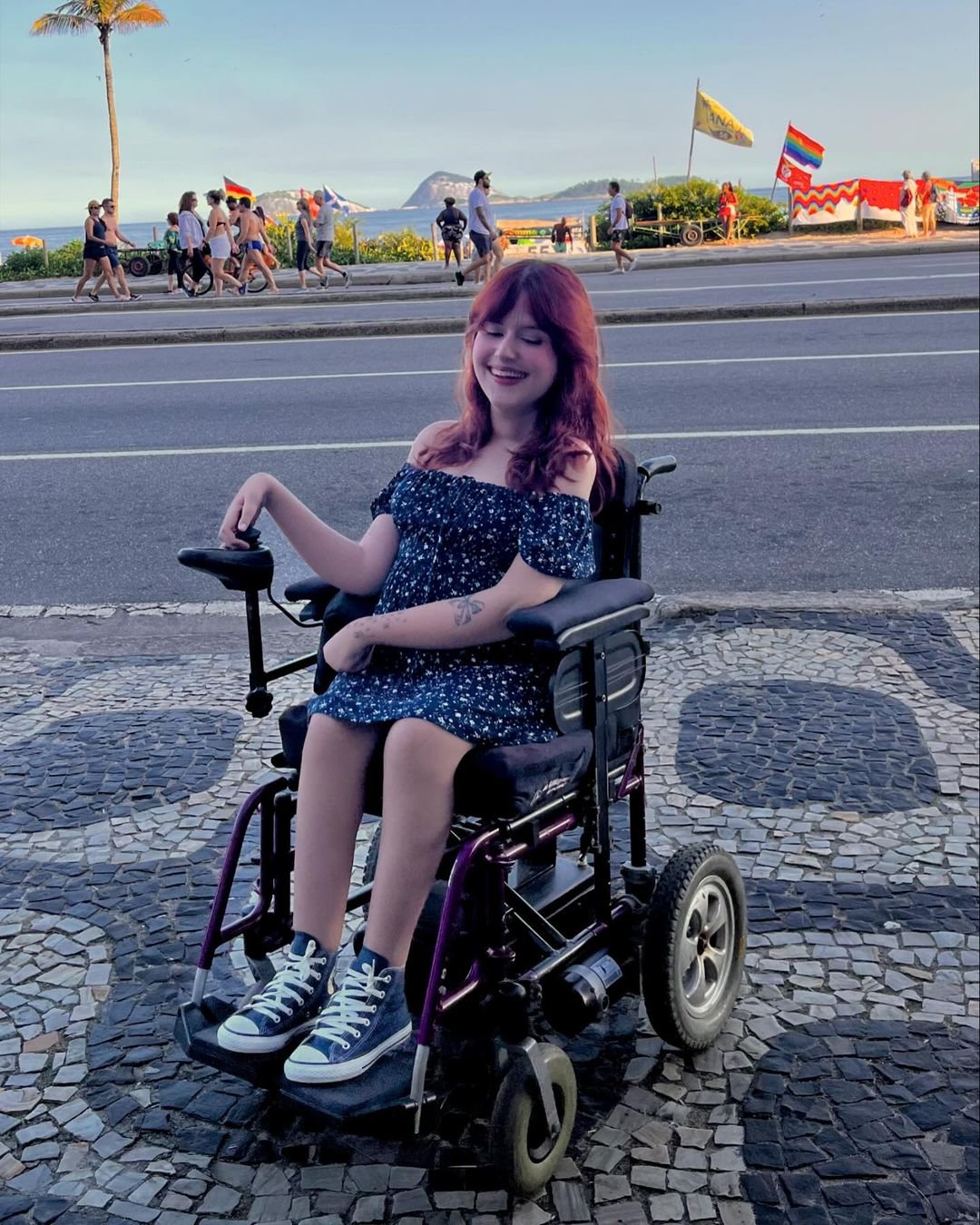 Pessoa em cadeira de rodas veste vestido floral azul e sapatos estilo Converse. O look casual, com vestido ombro a ombro, reflete tendências de conforto e moda despojada. Cabelos ruivos soltos complementam o visual jovem e descontraído. Marina Melo