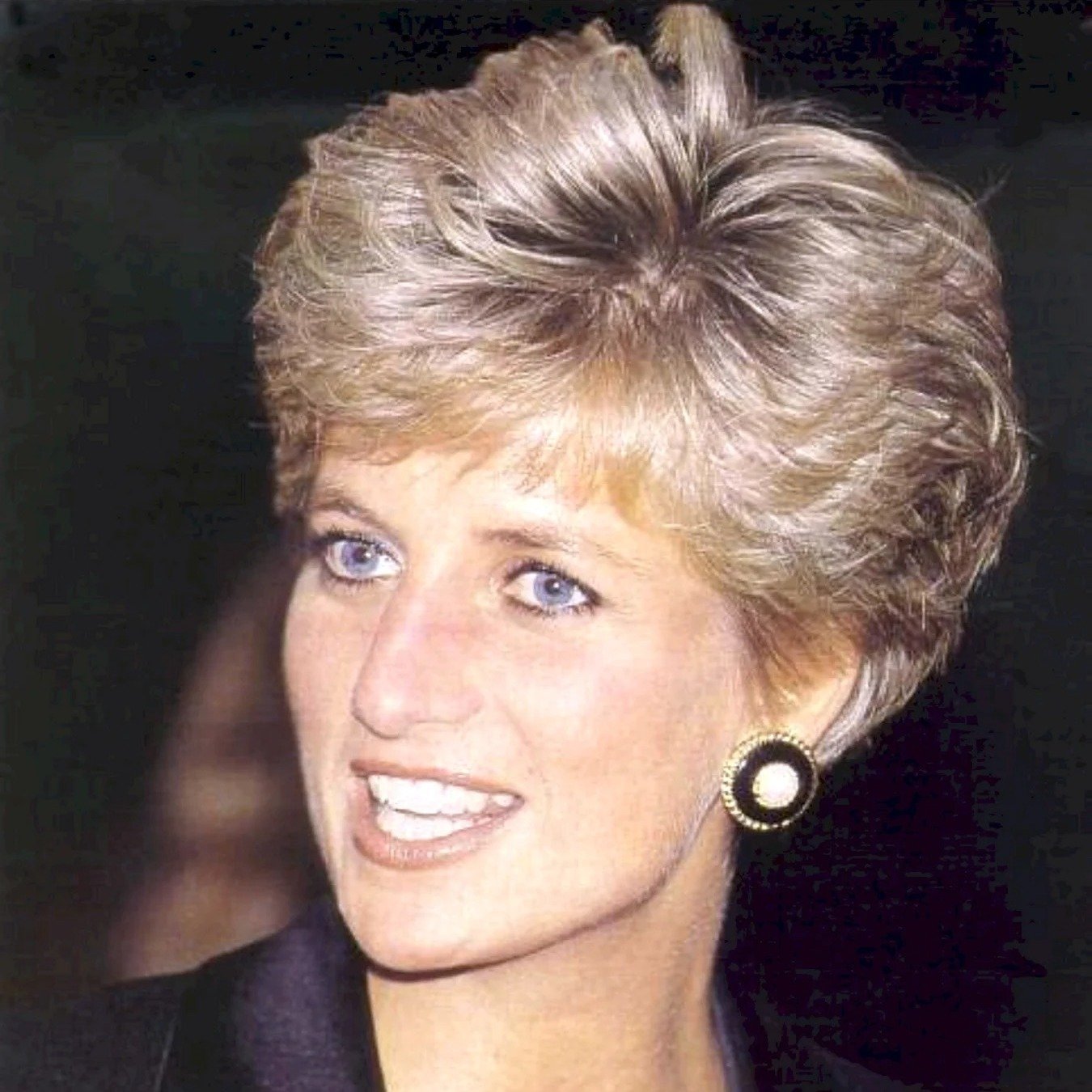 Imagem de uma mulher com cabelo loiro curto e volumoso no estilo anos 80/90. Ela usa brincos grandes e redondos com detalhes em dourado e pérola. A vestimenta inclui uma jaqueta preta clássica. Visual elegante e atemporal.