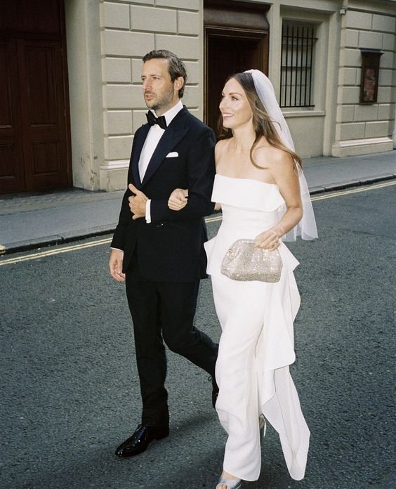 Casal elegante em cerimônia de casamento. Homem usa um clássico smoking preto, enquanto a mulher opta por um moderno macacão branco, com véu e clutch dourada. Tendência minimalista e sofisticada, destacando a inovação no vestuário nupcial.