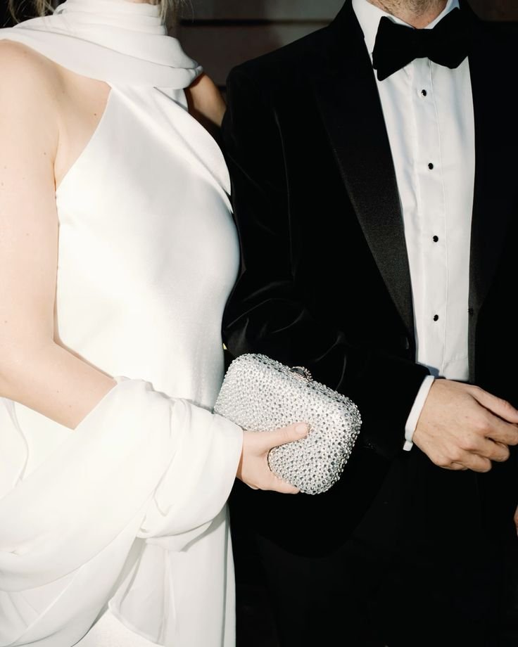 Casal elegante usa trajes formais: mulher em vestido branco de cetim com bolsa de pedrarias, homem com smoking preto e gravata borboleta. Look sofisticado e clássico, ideal para eventos de gala e festas de luxo.