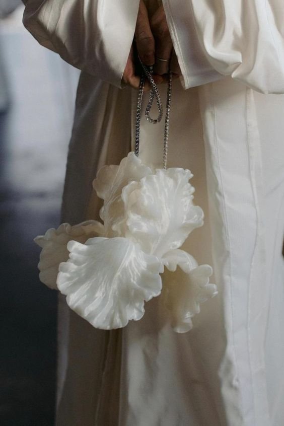 Pessoa vestindo traje longo e fluido de cor clara. A moda destaca detalhes elegantes e minimalistas, acompanhada de acessório único, uma bolsa escultural em formato de flores na cor branca, com alças de corrente metálica, representando tendência moderna e artística.