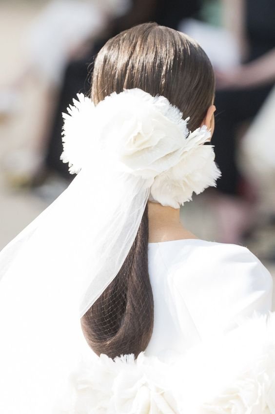 Imagem de costas mostrando penteado sofisticado com cabelo liso preso, decorado com grande laço floral branco e véu fino. Tendência de moda em acessórios de cabelo para noivas, destacando elegância e delicadeza para cerimônias contemporâneas.