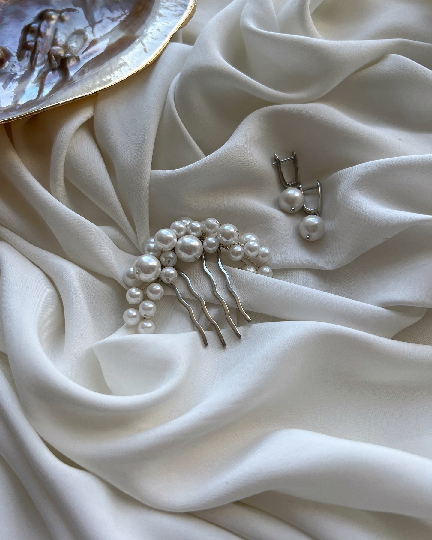 A imagem mostra acessórios elegantes como brincos de pérolas e um pente decorativo com pérolas, dispostos sobre tecido branco delicado, sugerindo um estilo clássico e sofisticado. Tendência contemporânea em moda nupcial e eventos formais.