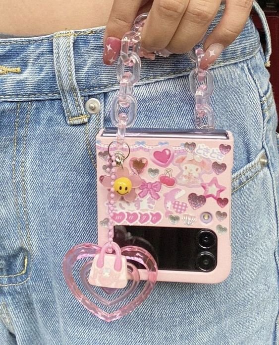 Celular dobrável com capa decorada com adesivos fofos e detalhes em rosa, pendurado em uma corrente de plástico. Tendência de moda kawaii, complementado por calça jeans de cintura alta, destacando o estilo jovem e divertido.