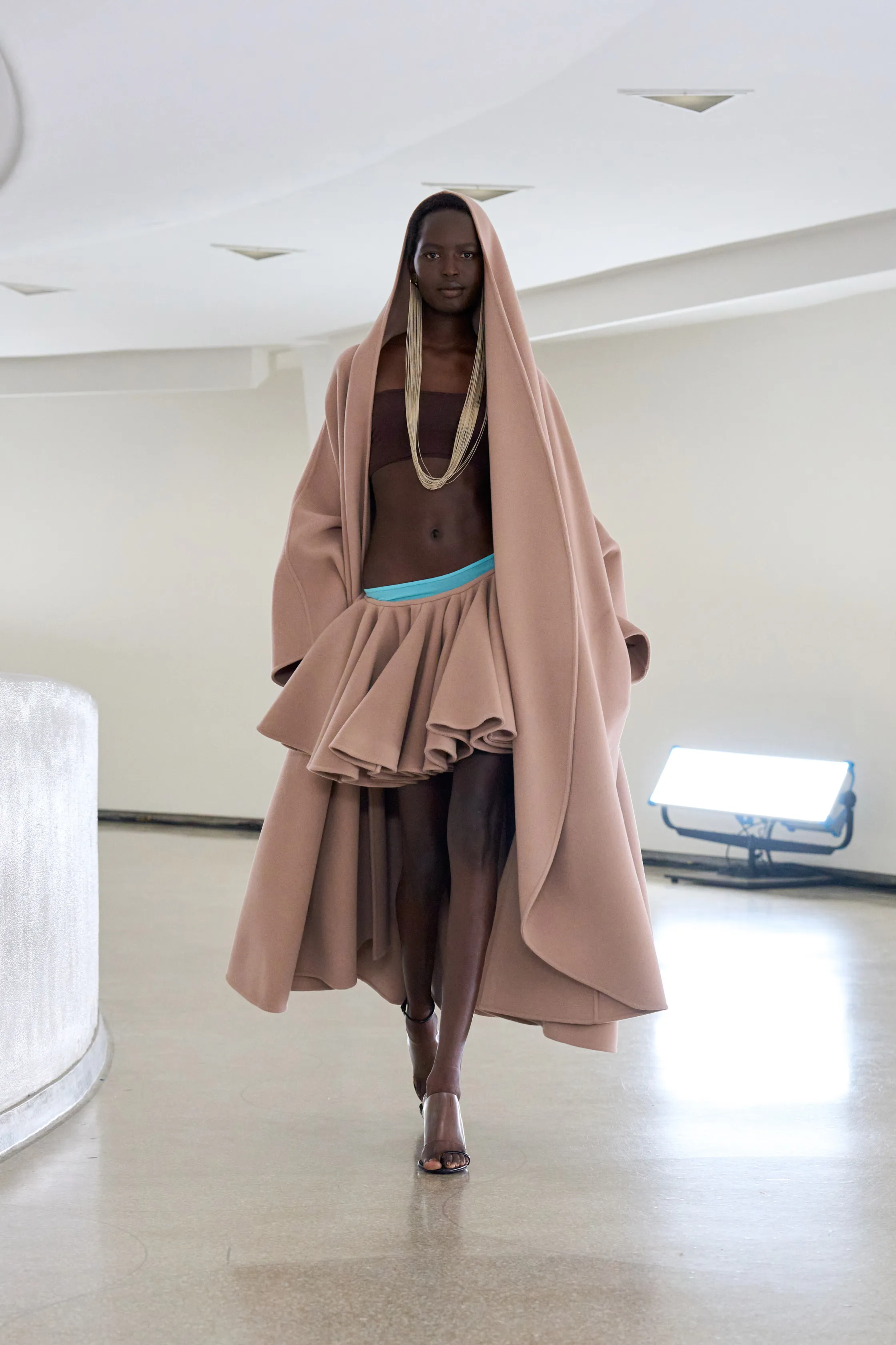 Modelo desfilando com tendências modernas de moda: capa oversized em tom nude, saia volumosa com babados e top minimalista. Acessórios incluem um colar longo e sandálias de salto fino, destacando uma estética inovadora e elegante.