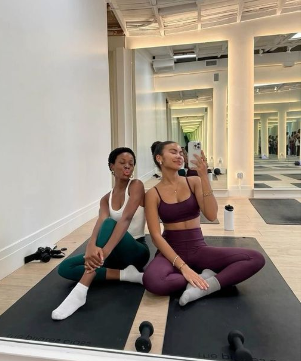 Duas mulheres vestindo roupas de academia posam para foto. Uma delas usa top branco e calça legging verde escura, enquanto a outra usa conjunto de top e calça legging roxa. Ambos os looks são tendências no segmento fitness, destacando conforto e estilo.