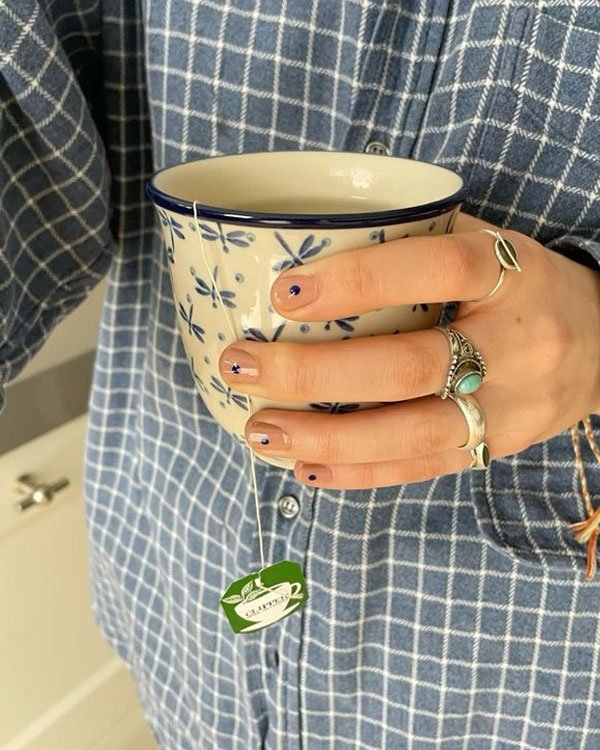 Pessoa usando camisa xadrez azul e segurando xícara estampada. Unhas decoradas com pequenos desenhos e vários anéis estilosos complementam o visual moderno e casual.