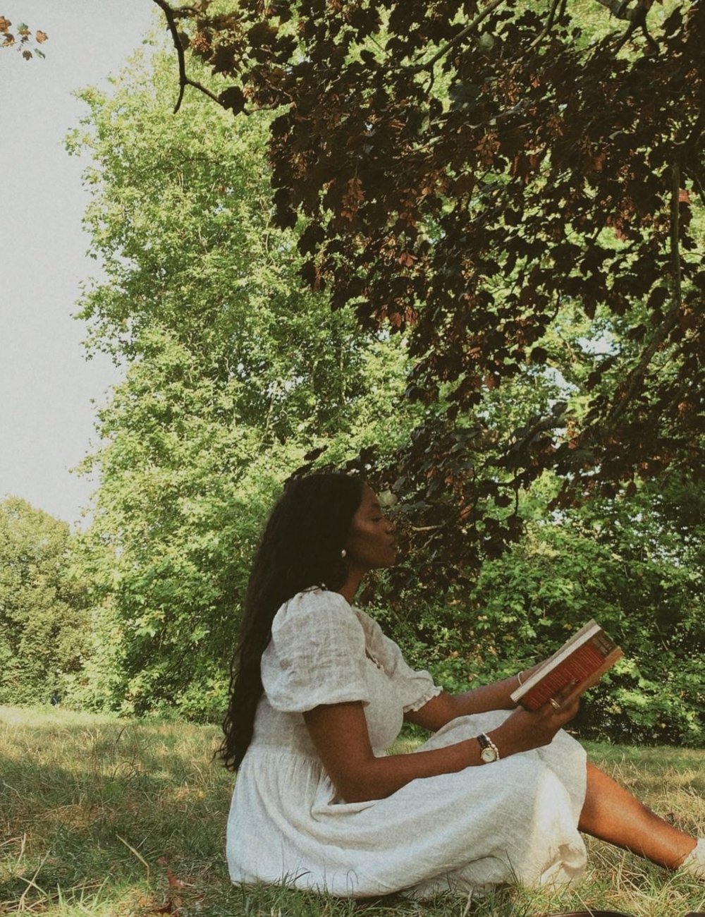 Mulher sentada ao ar livre lendo um livro, veste um vestido branco estilo cottagecore, de mangas bufantes e tecido leve. Tendência romântica e vintage, ideal para dias ensolarados. Ansiosa sobre o futuro.